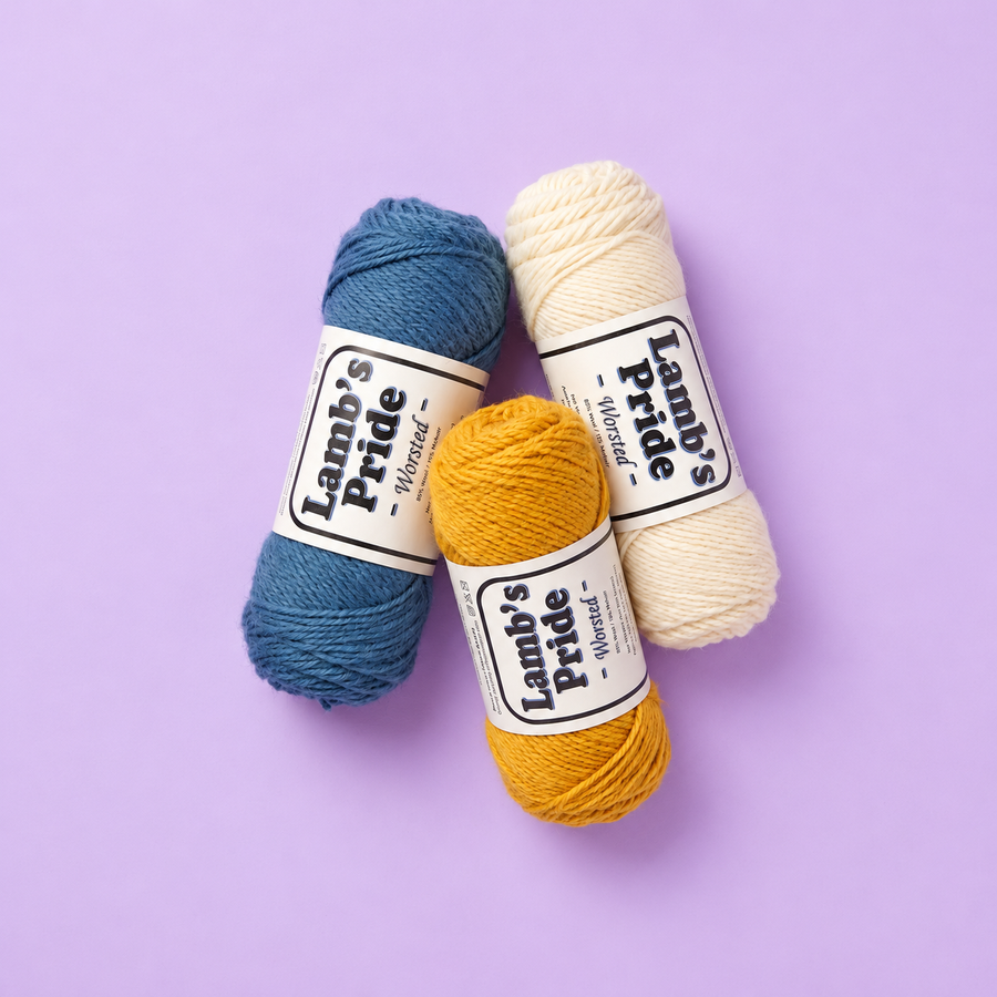 Three Brown Sheep Lamb’s Pride Worsted yarn skeins in blue, cream, and gold with paper labels on a soft lavender background.