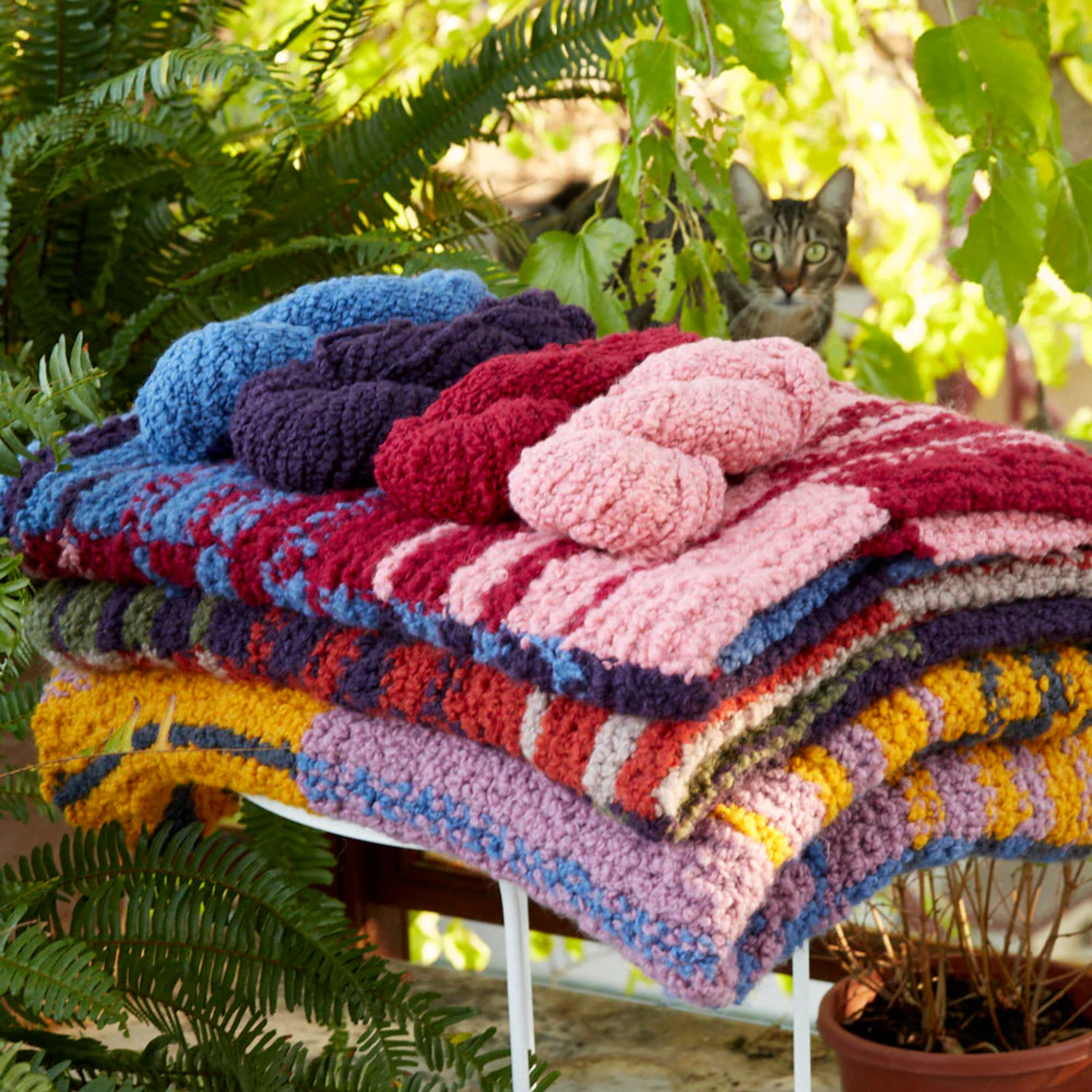 Stack of three Accoloration Blankets in Twilight Harvest, Golden Hour, and Crimson Sky colorways. Four skeins of yarn in opal, amethyst, quartz, and garnet from the Crimson Sky variant are arranged on top. A cat peeks out from behind the stack, surrounded by greenery.