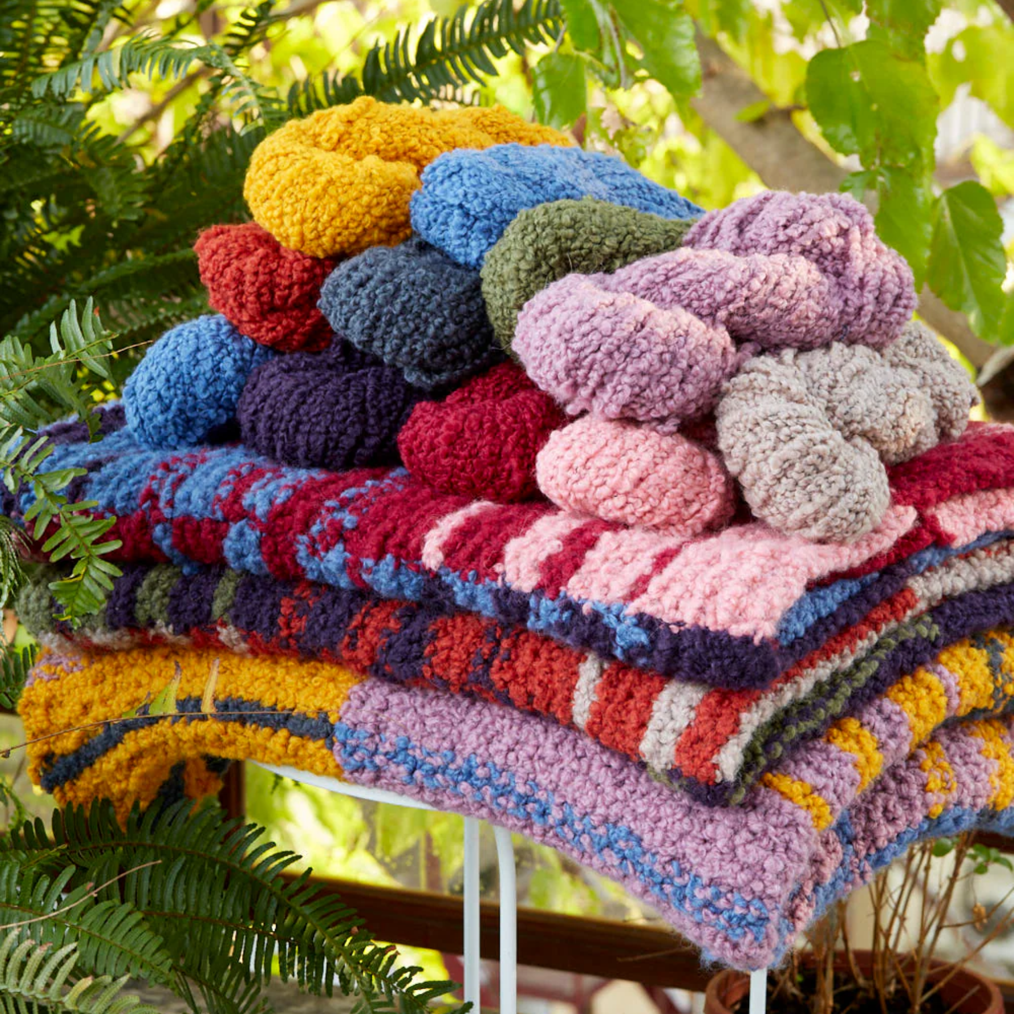 Stack of three Accoloration Blanket Kits with coordinating skeins of yarn in warm, vibrant colors. The blankets feature bold stripes and gradient color transitions, showcasing the Twilight Harvest, Golden Hour, and Crimson Sky variants.