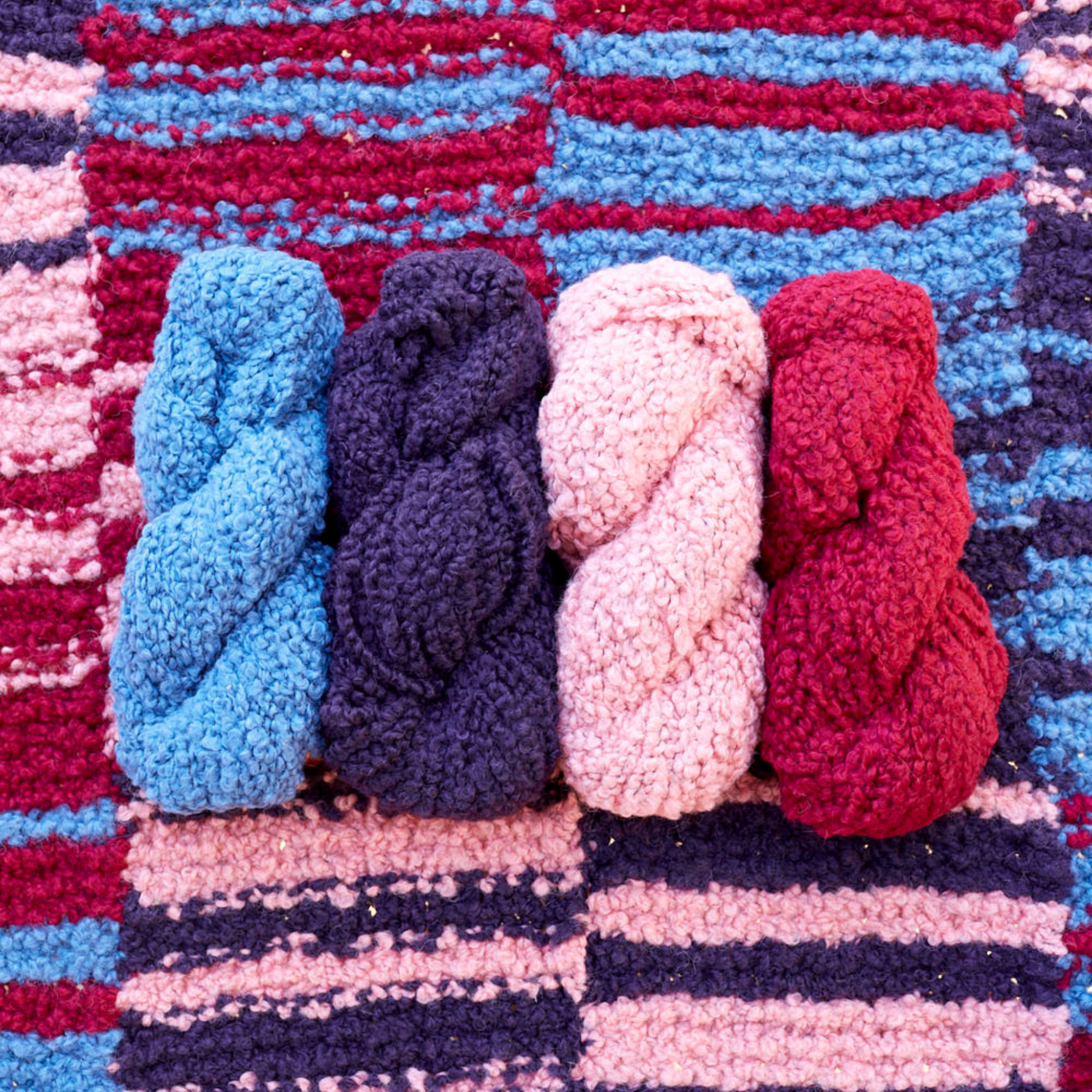 Four skeins of Lanalpaca yarn in opal, amethyst, quartz, and garnet colors arranged on the Crimson Sky Accoloration Blanket, showcasing the rich reds, pinks, and blues in the striped design.