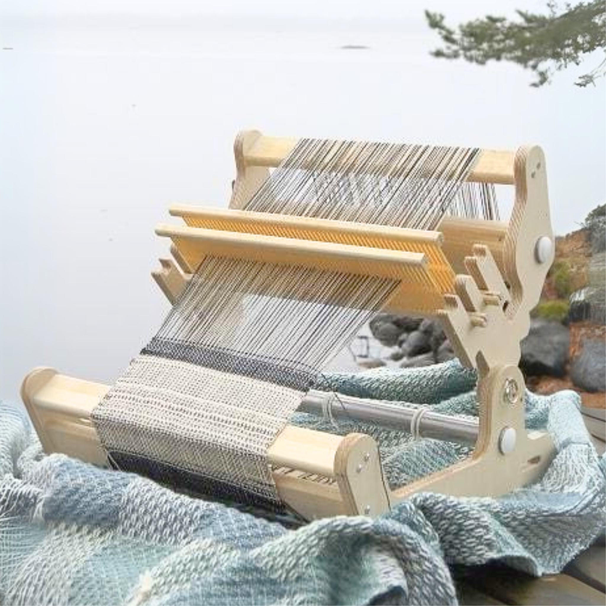 Glimakra Siru Rigid Heddle Loom set up outdoors by a lakeside, showcasing its portability and ease of use for weaving on the go. The ergonomic design and smooth weaving action make it a perfect choice for travel-friendly fiber arts.