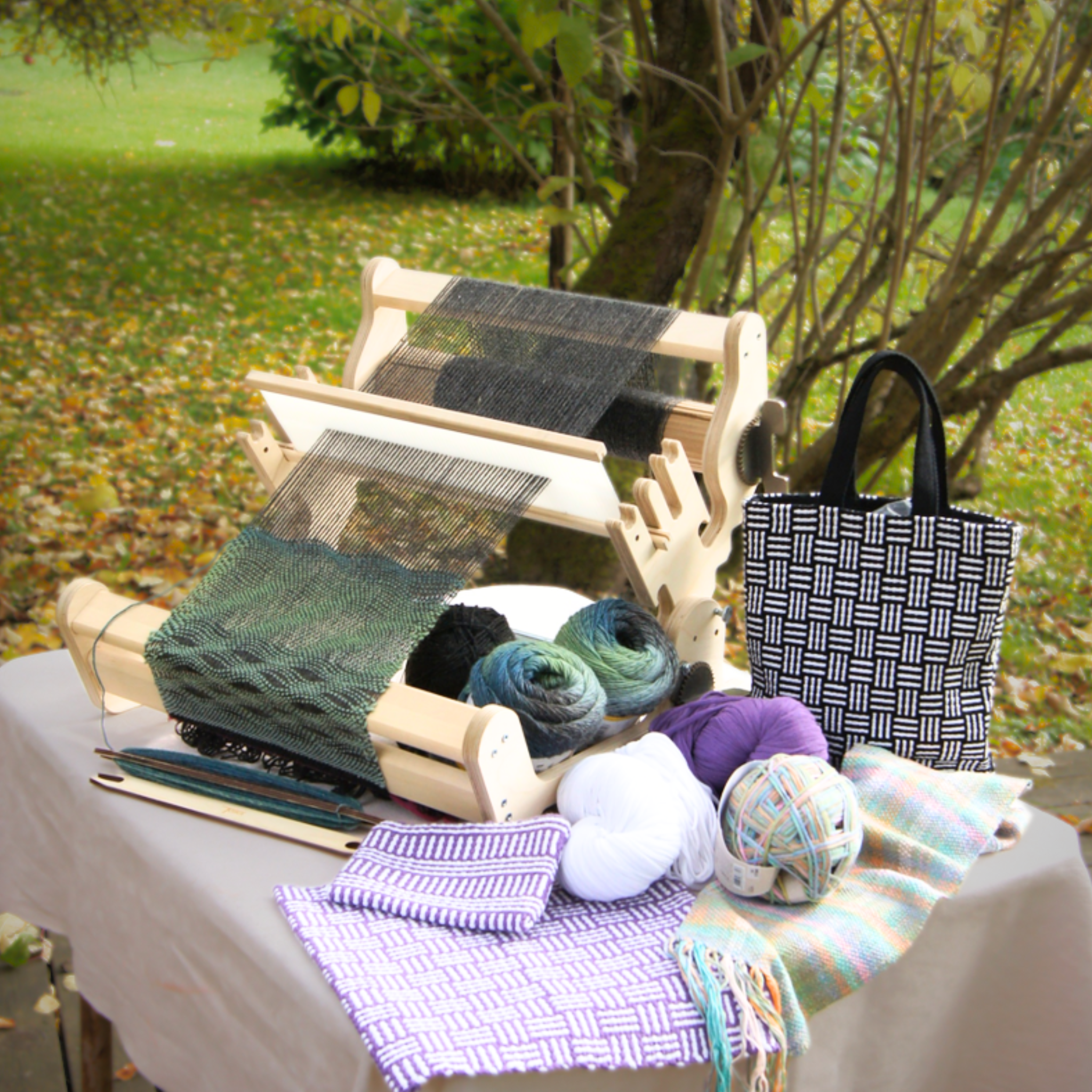 Glimakra Siru Rigid Heddle Loom set up outdoors with various weaving projects, yarn skeins, and woven textiles. This ergonomic loom is perfect for portable weaving, offering versatility for different yarns and patterns, ideal for both beginner and advanced weavers.