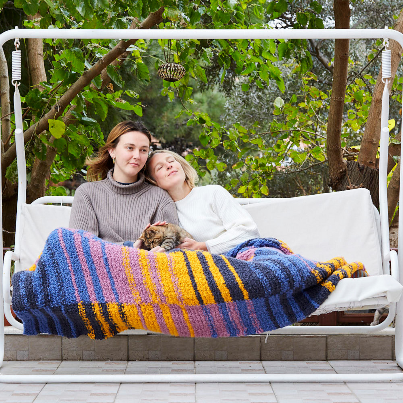 Two people sitting on an outdoor swing, wrapped in the Golden Hour Accoloration Blanket featuring bold stripes of blue, gold, and purple. A cat rests on their lap as they relax under the trees.