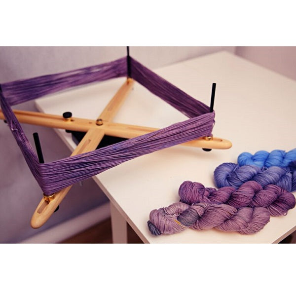 Clear Finish Kromski Vivo Skein Winder in use with purple yarn being wound into a skein, shown alongside gradient skeins in blue and purple tones. Perfect for spinners, weavers, and knitters creating organized skeins with ease and precision.