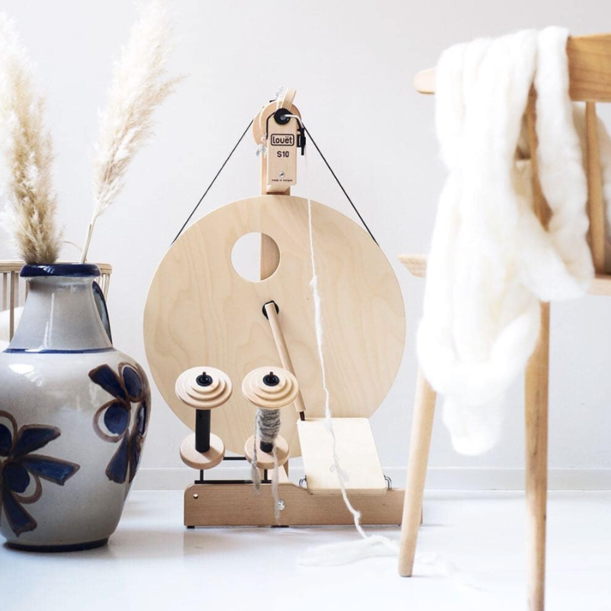 Lifestyle image of Louet S10 Single Treadle Spinning Wheel styled with ceramic vase, roving, and wooden chair, illustrating a cozy fiber art workspace.