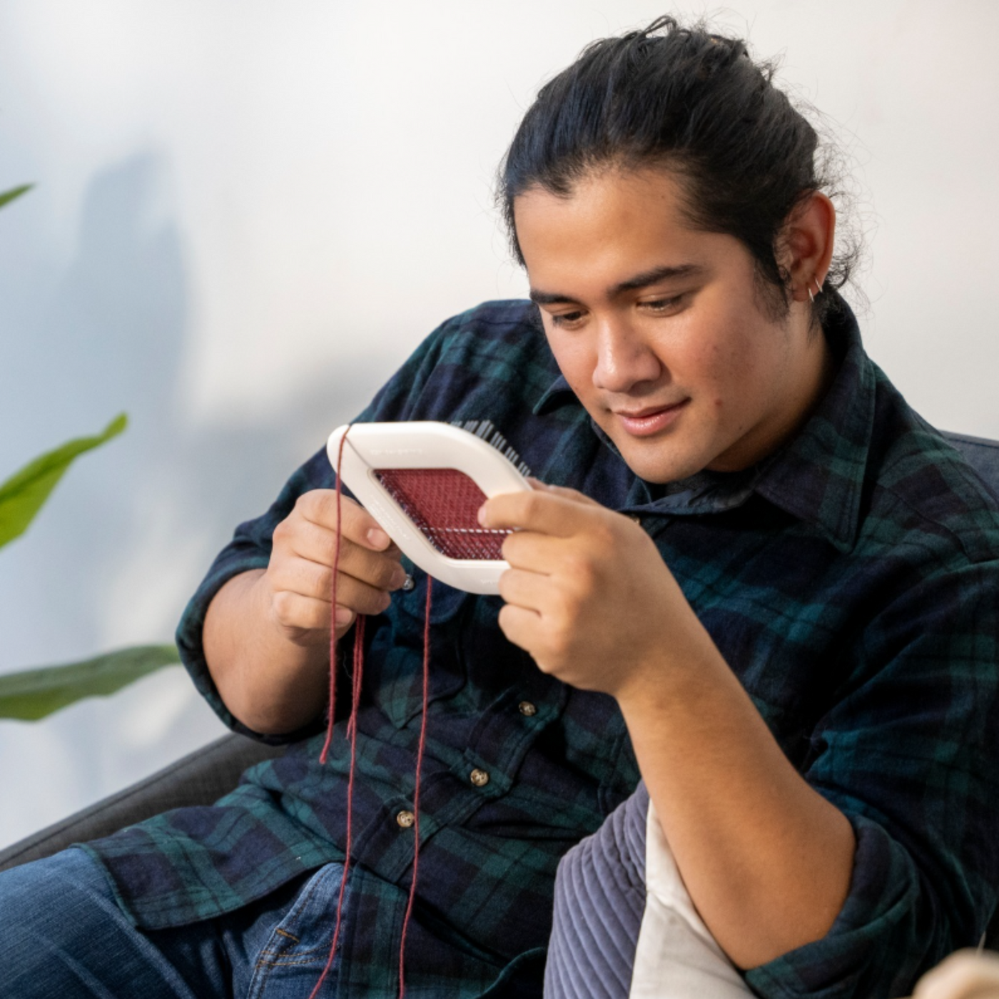 A man in a plaid shirt weaving on the Schacht Zoom Loom, a portable pin loom for creating woven squares. Seated comfortably indoors, he focuses on his weaving project with red yarn.