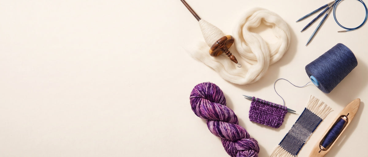 Purple yarn, undyed wool fiber, knitting needles, drop spindle, indigo yarn cone, and weaving shuttle on a warm cream background.