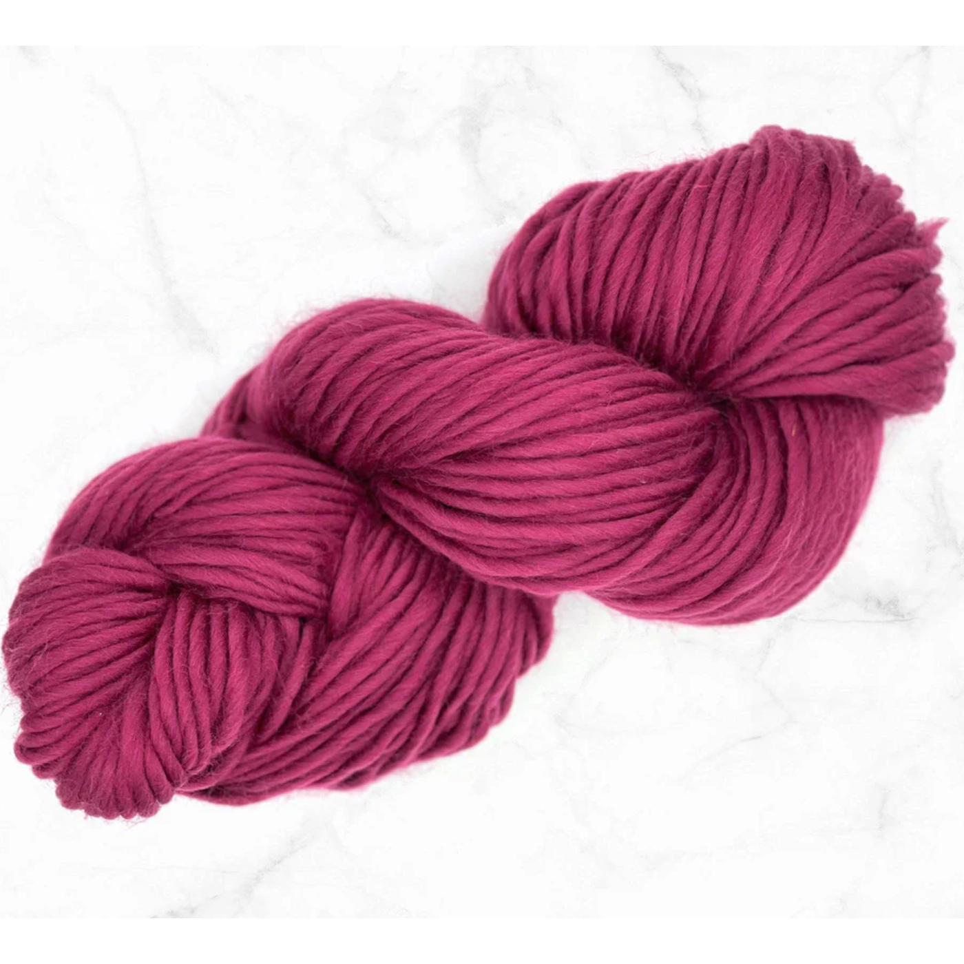 A skein of super bulky weight merino yarn in the color mulberry, a rich cool tone reddish purple. Placed on a white marble background.