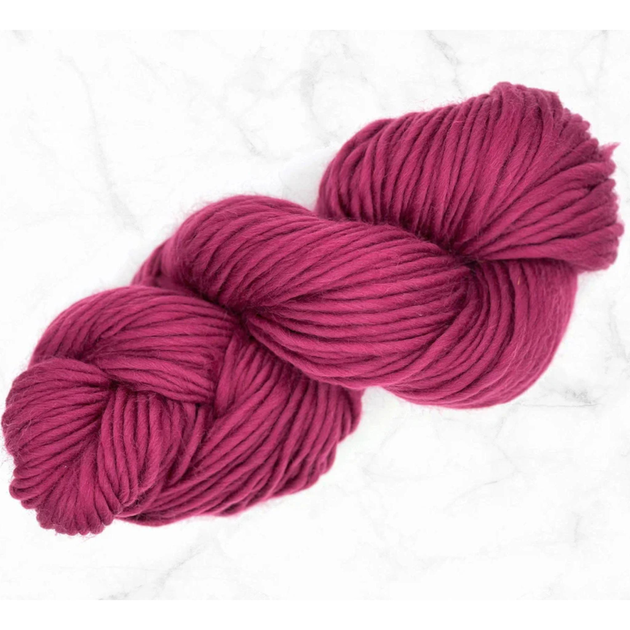 A skein of super bulky weight merino yarn in the color mulberry, a rich cool tone reddish purple. Placed on a white marble background.