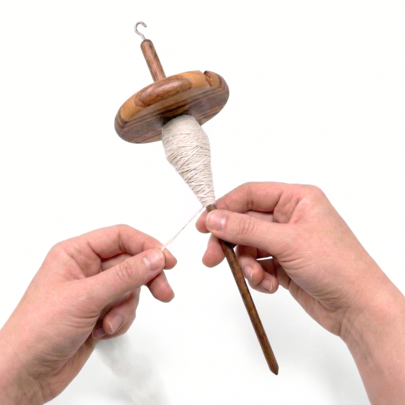 Hands drafting fiber while spinning with a Revolution Fibers top whorl drop spindle; white singles on the spindle, clean white background