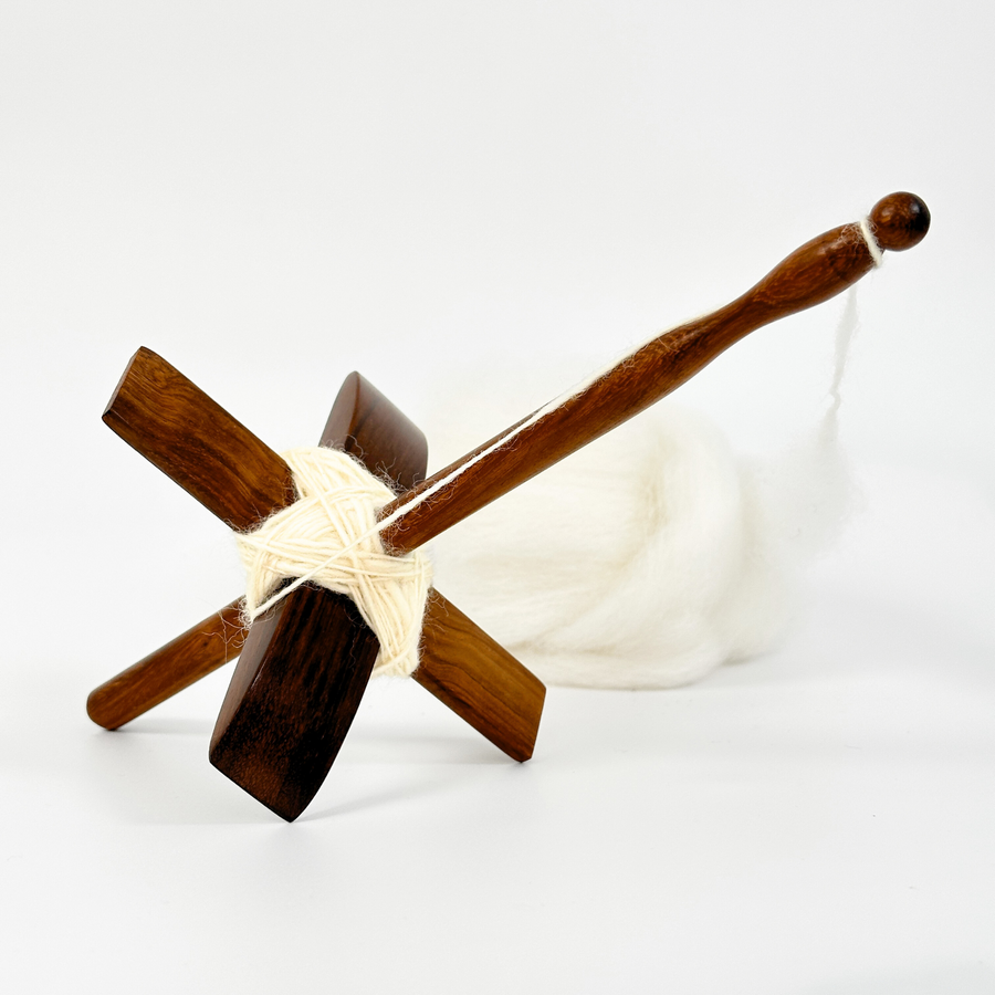 Angled studio view of Turkish drop spindle resting beside cream wool roving and spun yarn, showcasing a portable spinning tool in use.