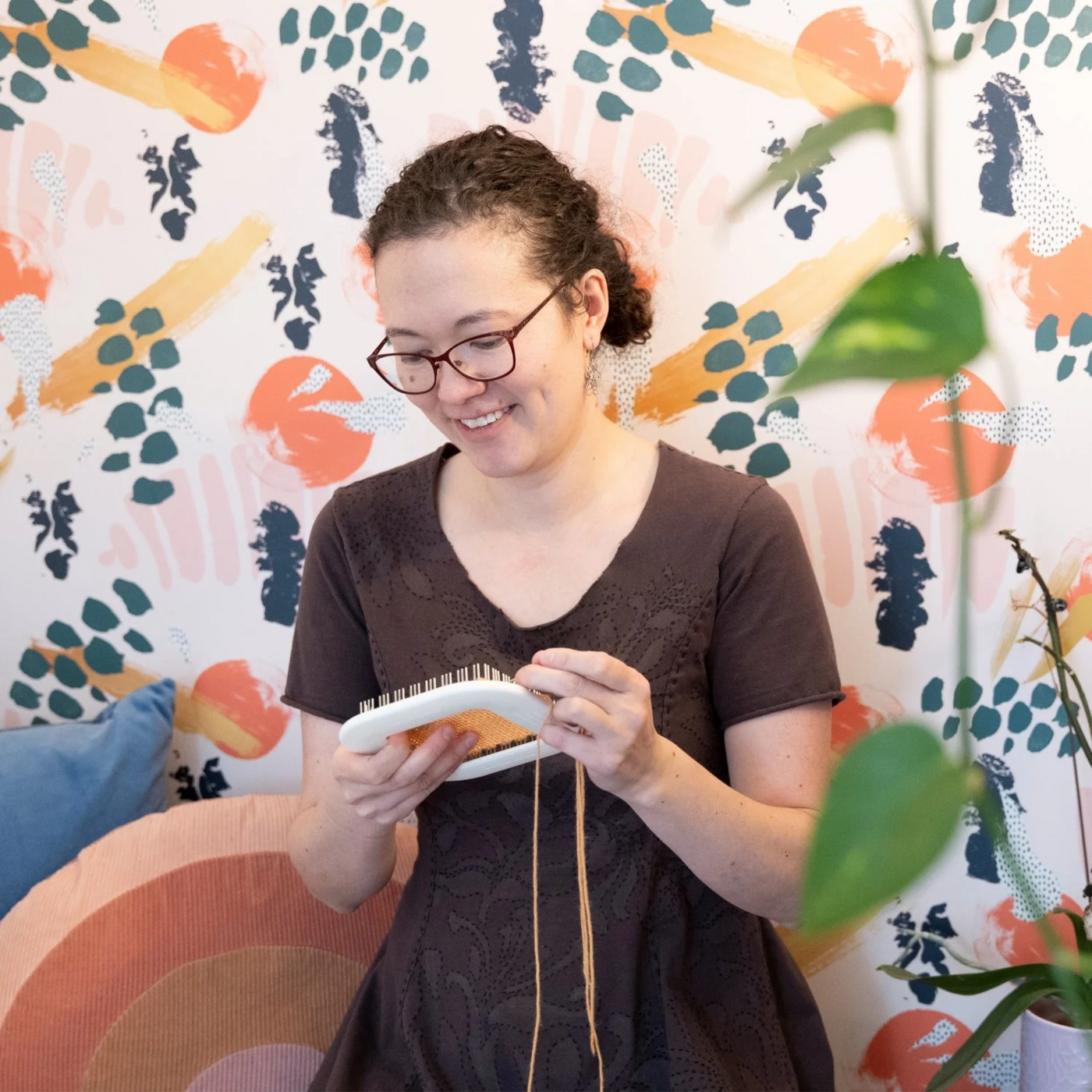 A smiling woman weaving on the Schacht Zoom Loom, a portable 4x4 pin loom for crafting handmade textiles, small woven squares, and creative fiber art projects.