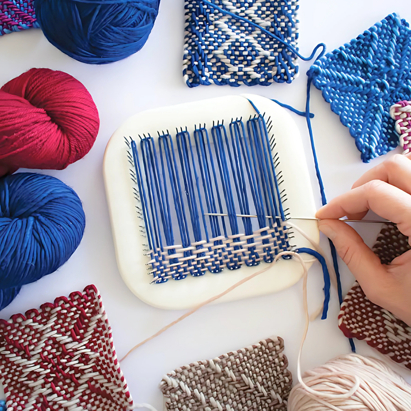 A close-up of the Schacht Zoom Loom with blue yarn in progress, surrounded by handwoven pin loom squares and skeins of red, blue, and beige yarn, showcasing intricate weaving techniques.