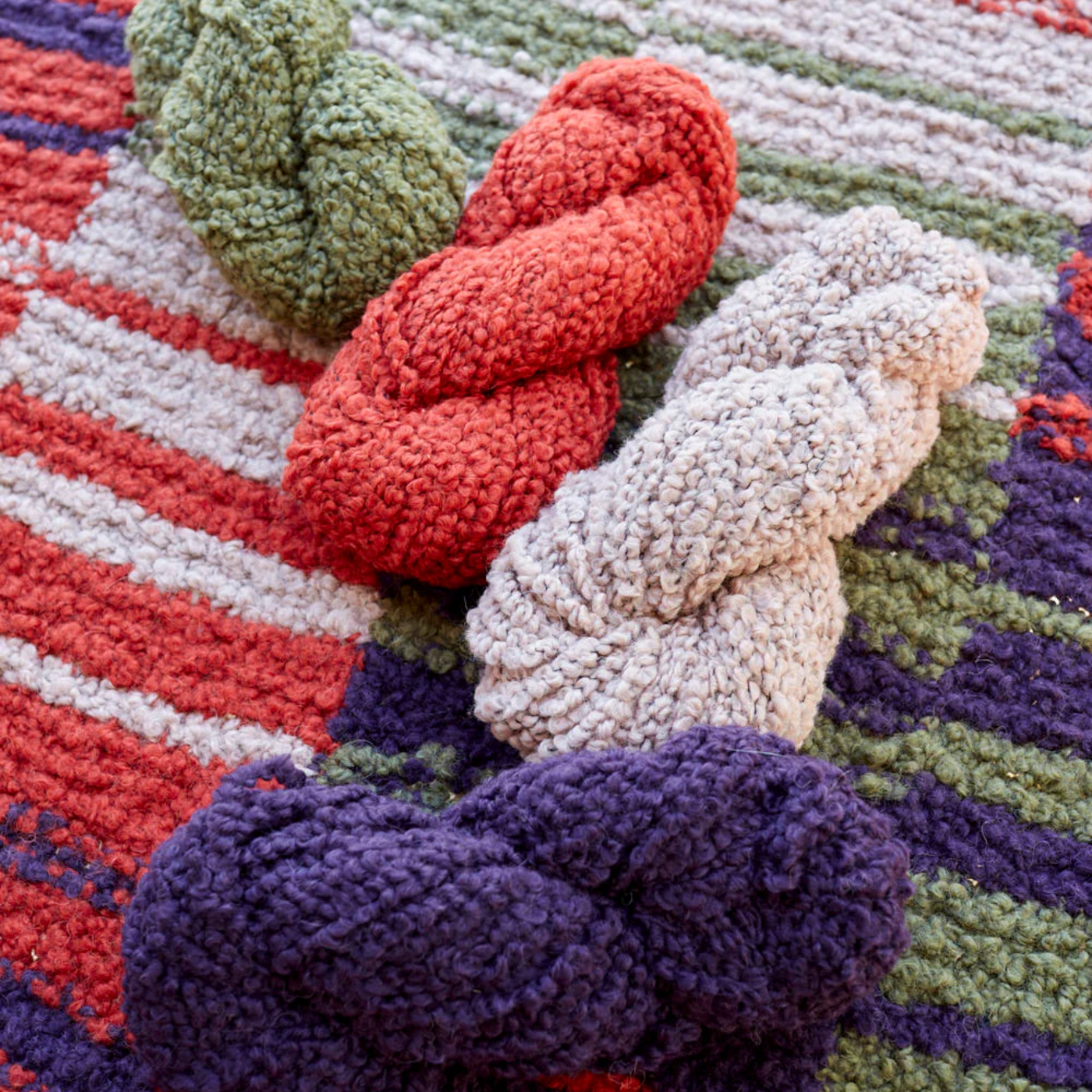 Four skeins of Lanalpaca yarn in amethyst, travertine, copper, and pumice colors, arranged on top of the Twilight Harvest Accoloration Blanket featuring matching striped and block patterns.