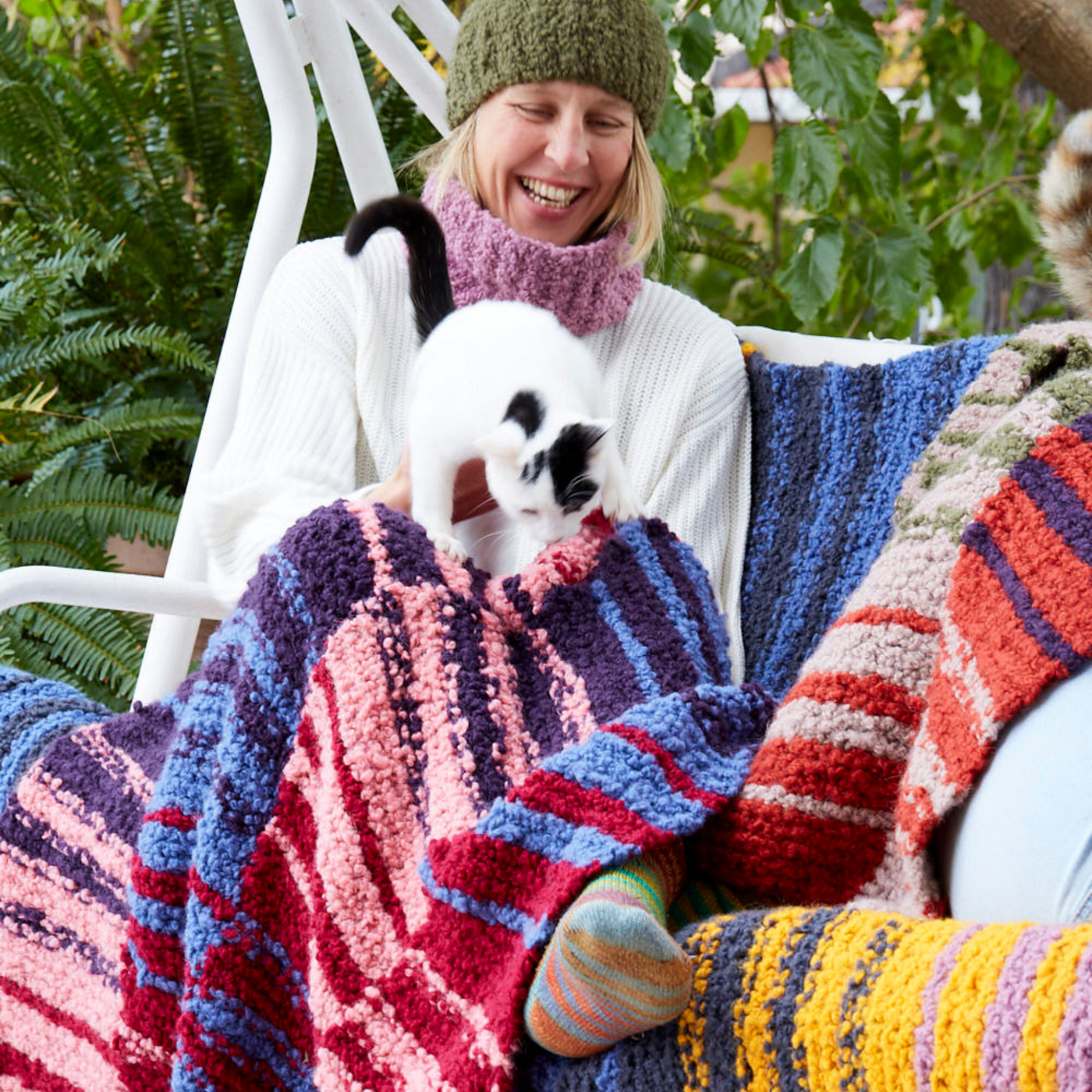 Person sitting on an outdoor swing, smiling as a black and white cat walks across the Crimson Sky Accoloration Blanket. The Twilight Harvest and Golden Hour blanket variants are also draped over the swing, showcasing all three colorways.