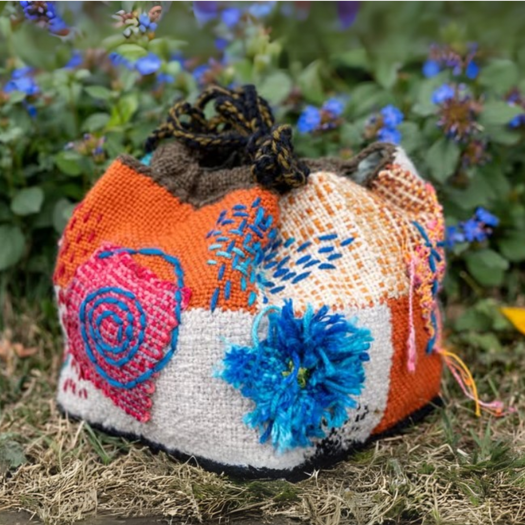 A colorful, textured handwoven bag made from pin loom squares, featuring embroidery, tufted yarn embellishments, and bold patterns, created using the Schacht Zoom Loom. Displayed outdoors against a floral background.